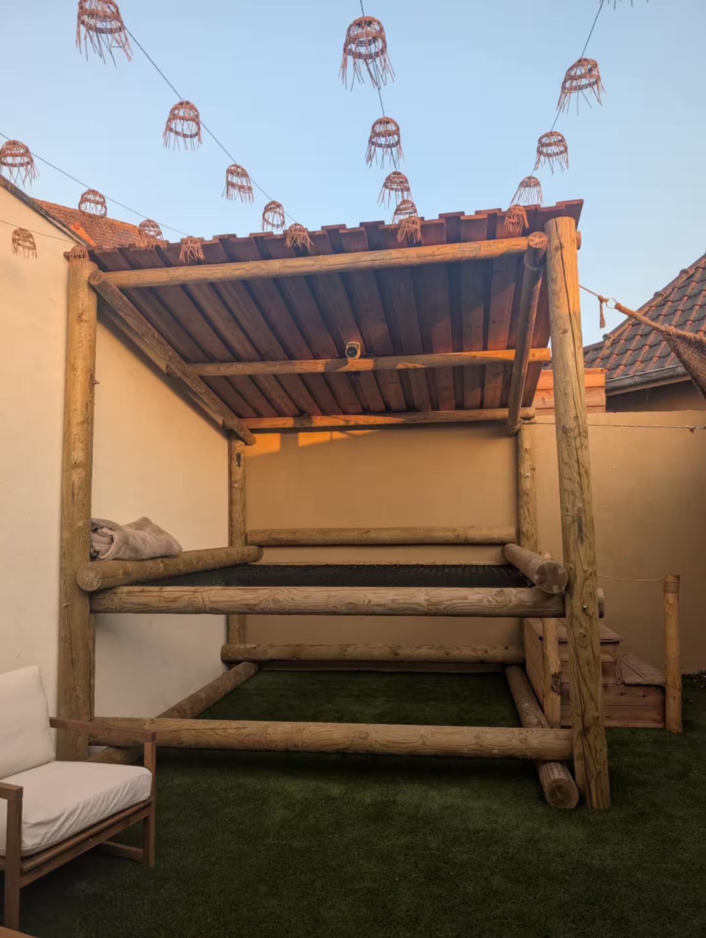 Lit de repos en rondins sous une pergola en bois, guirlande de lanternes en osier au coucher du soleil